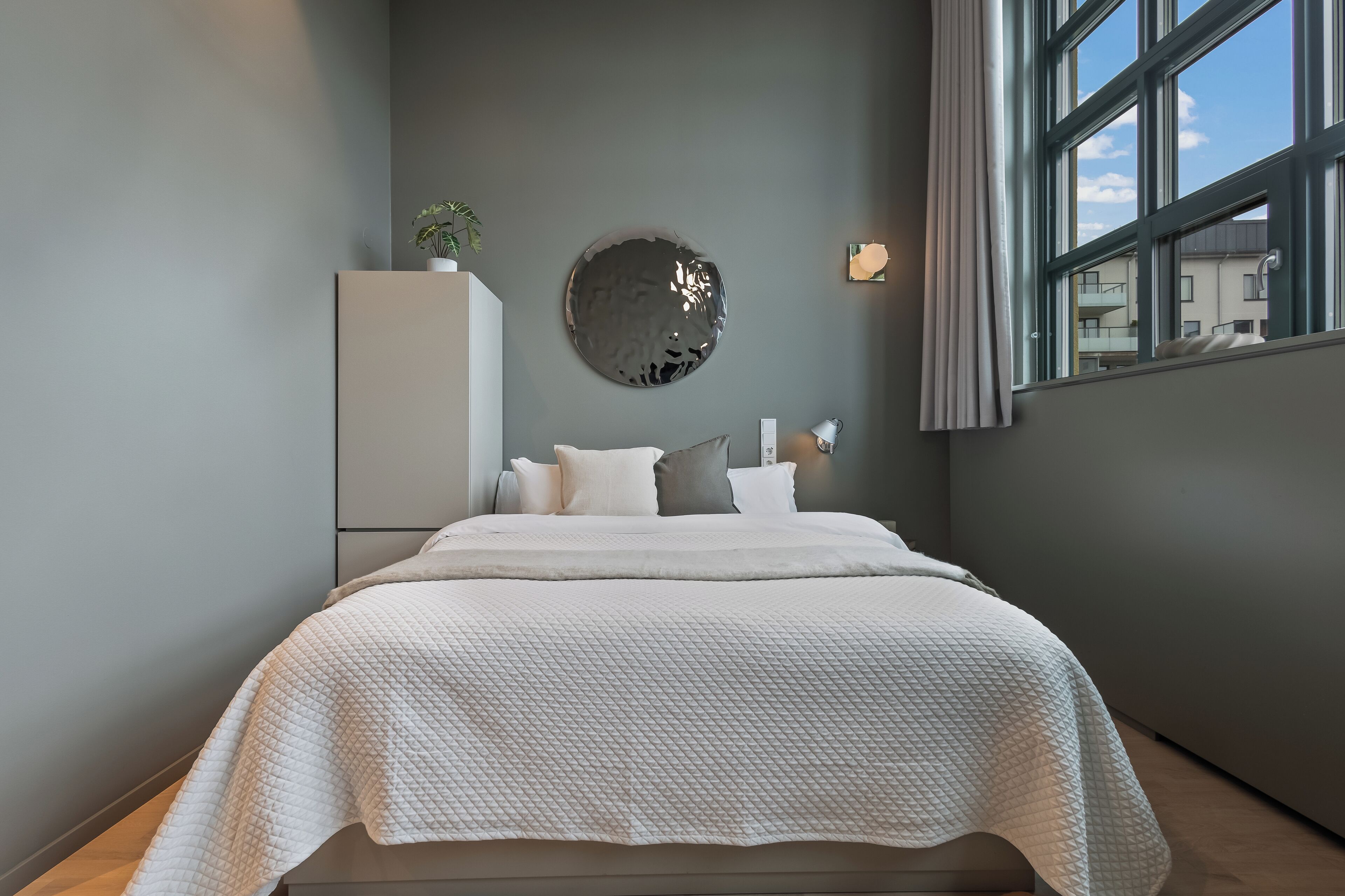A modern bedroom with a large bed, a wall-mounted mirror, and a view of the outside through a window.