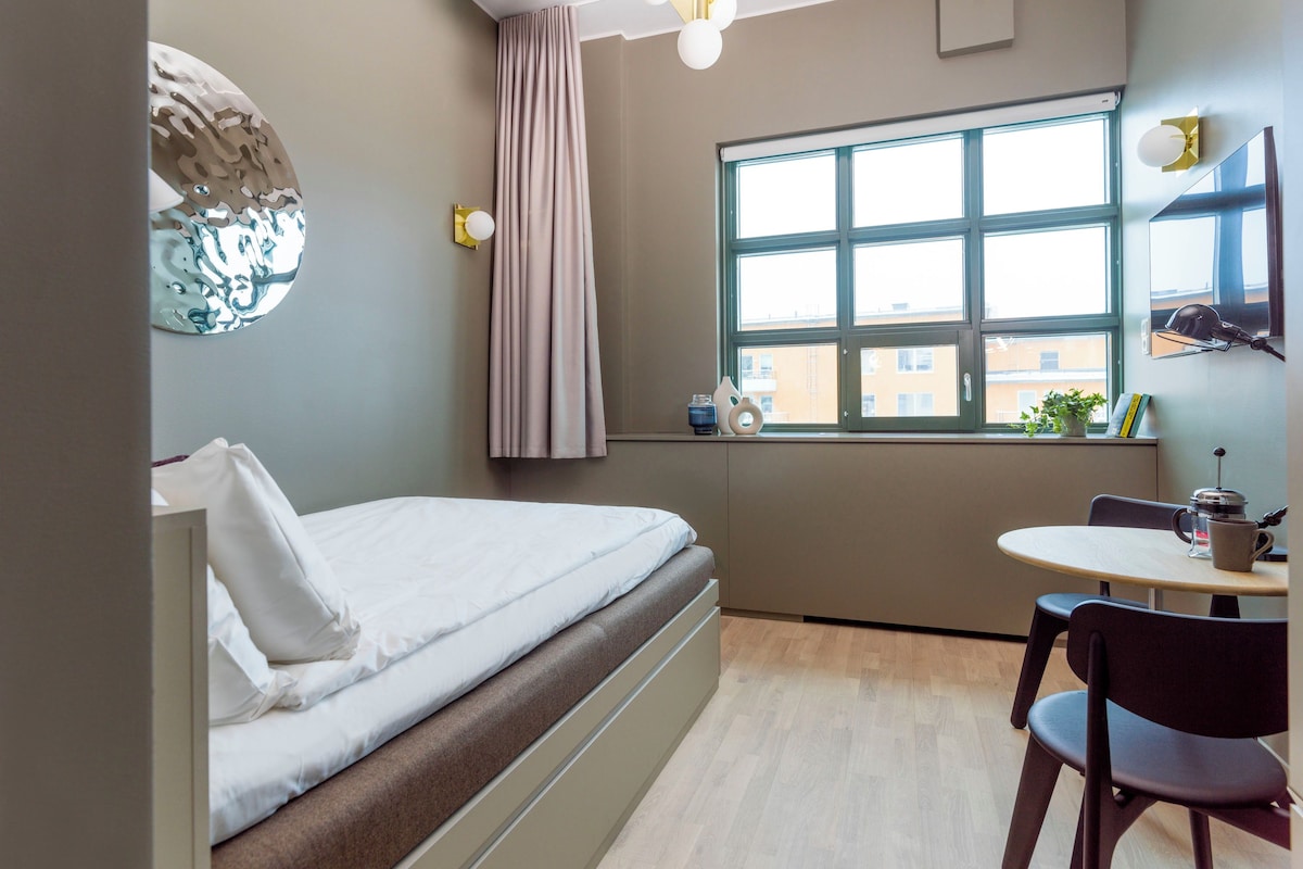 A compact hotel room with a bed, a small table, a chair, a window with a view, and a ceiling light fixture.