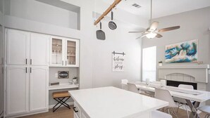A modern kitchen with white cabinetry, a central island, and a dining area with white chairs. There is a ceiling fan, a painting on the wall, and a sign that reads "Welcome to our story". - 35% off Special Green Valley Gem! 3/2 Single Story (Henderson)
