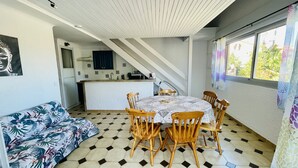 En matplats med ett runt bord och trästolar, en soffa med ett tropiskt mönster och ett kök i bakgrunden.