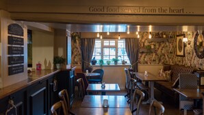 A cozy restaurant interior with a wooden counter, multiple seating options including tables and a sofa, and a window with curtains. - The Brewers Inn (Cambridge)
