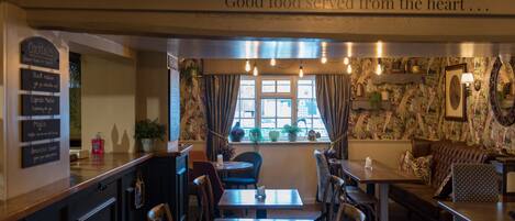 Un acogedor interior de restaurante con mostrador de madera, diversas opciones de asientos, incluyendo mesas y un sofá, y una ventana con cortinas.