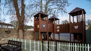 A wooden play structure with a white picket fence, a bench, and a tree in the background. - The Brewers Inn (Cambridge)