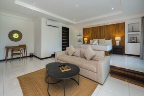 Une chambre d’hôtel moderne, équipée d’un canapé, d’une petite table, d’un lit avec une tête de lit en bois et d’une table de chevet avec une lampe.