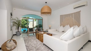 A modern living room with a white sofa, a coffee table, and a wicker chair. There is a large window with a view of a pool and greenery outside. - Paradise Beach Villa Santa Pila (San Miguel de Cozumel)