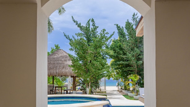 A view through an arched window of a pool area with a thatched-roof structure, lounge chairs, and a clear sky. - Paradise Beach Villa Santa Pila (San Miguel de Cozumel)
