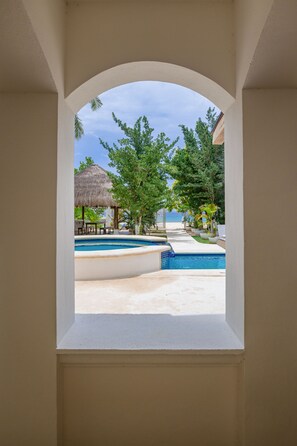 A view through an arched window of a pool area with a thatched-roof structure, lounge chairs, and a clear sky. - Paradise Beach Villa Santa Pila (San Miguel de Cozumel)