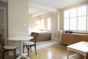 A room with a bed, a dining table, chairs, a small kitchenette, and a window with blinds.