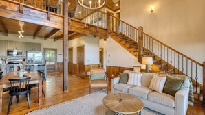 A spacious living area with a wooden staircase, a dining table with chairs, and a cozy seating area with a sofa and cushions. - Fab-VIEW-lous 4-bdrm cabin w/ Hottub, LUX theater, outdoor fireplace & firepit (Morganton)