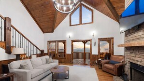 A spacious living room with a high ceiling, wooden beams, a fireplace, and large windows offering a view of the outdoors. - Fab-VIEW-lous 4-bdrm cabin w/ Hottub, LUX theater, outdoor fireplace & firepit (Morganton)