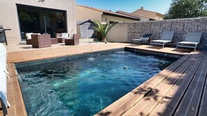 A swimming pool with wooden deck and lounge chairs, surrounded by a stone wall and a modern house. - Maison Paisible (VEDENE)