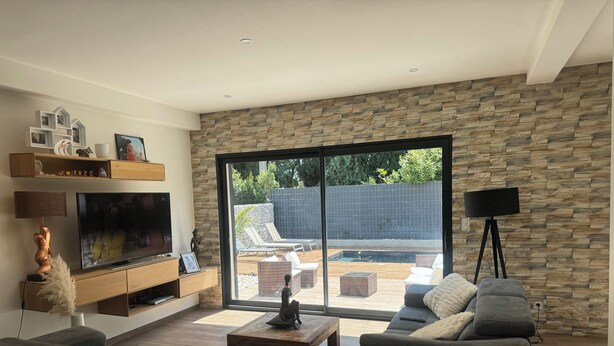 A modern living room with a stone wall, a large sliding glass door leading to a pool area, and a flat-screen TV on a wooden console. - Maison Paisible (VEDENE)