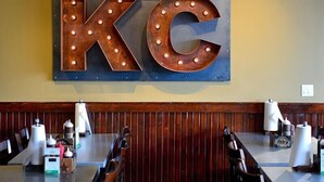 Ein Restaurantinneres mit einem großen, beleuchteten Schild, auf dem 'JOE'S KC' steht. Die Tische sind mit Gewürzen und Servietten gedeckt.