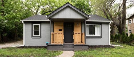 A small, single-story house with a gray exterior, a black front door, and a wooden porch with a railing.