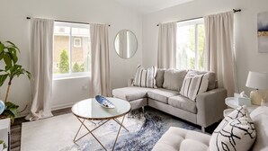 Ein Wohnzimmer mit einem beigefarbenen Sofa, einem runden Couchtisch und einem großen Fenster mit Vorhängen.