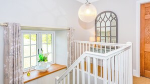 Un escalier avec des rampes blanches mène à un palier. On peut voir une fenêtre ornée de rideaux à motifs floraux et un vase rempli de fleurs sur le palier.