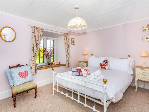 Un dormitorio con cama blanca, una silla de madera, una mesita de noche, una lámpara, un espejo y una ventana con cortinas.