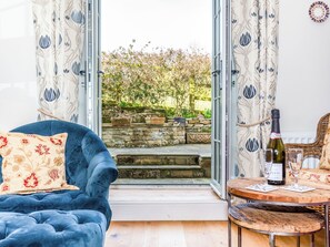 Una habitación con un sofá azul, una mesita de madera con una botella y vasos, y vista a un jardín a través de una puerta de vidrio.
