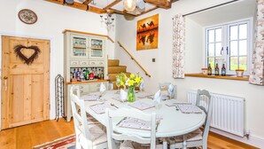 Un coin repas avec une table ronde dressée pour quatre personnes, entourée de chaises blanches dotées de coussins en tissu tressé. Un buffet en bois avec des portes en verre est visible sur la gauche, et un escalier avec une rampe en bois mène à l’étage supérieur.
