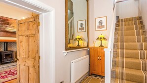 A hallway with a wooden door, a mirror, a radiator, a patterned rug, and a staircase with striped carpet. - The Farmhouse - UK33419 (West Witton, near Leyburn)
