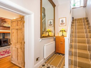 A hallway with a wooden door, a mirror, a radiator, a patterned rug, and a staircase with striped carpet. - The Farmhouse - UK33419 (West Witton, near Leyburn)