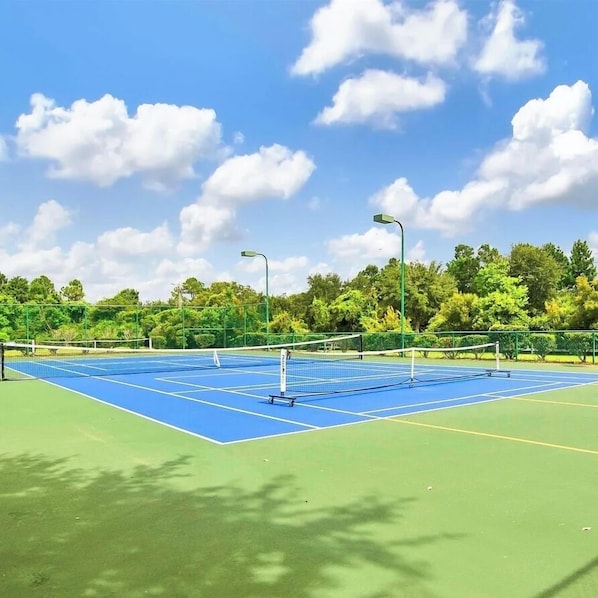 Un court de tennis en plein air avec une surface de jeu bleue, entouré d’arbres verdoyants et sous un ciel dégagé.