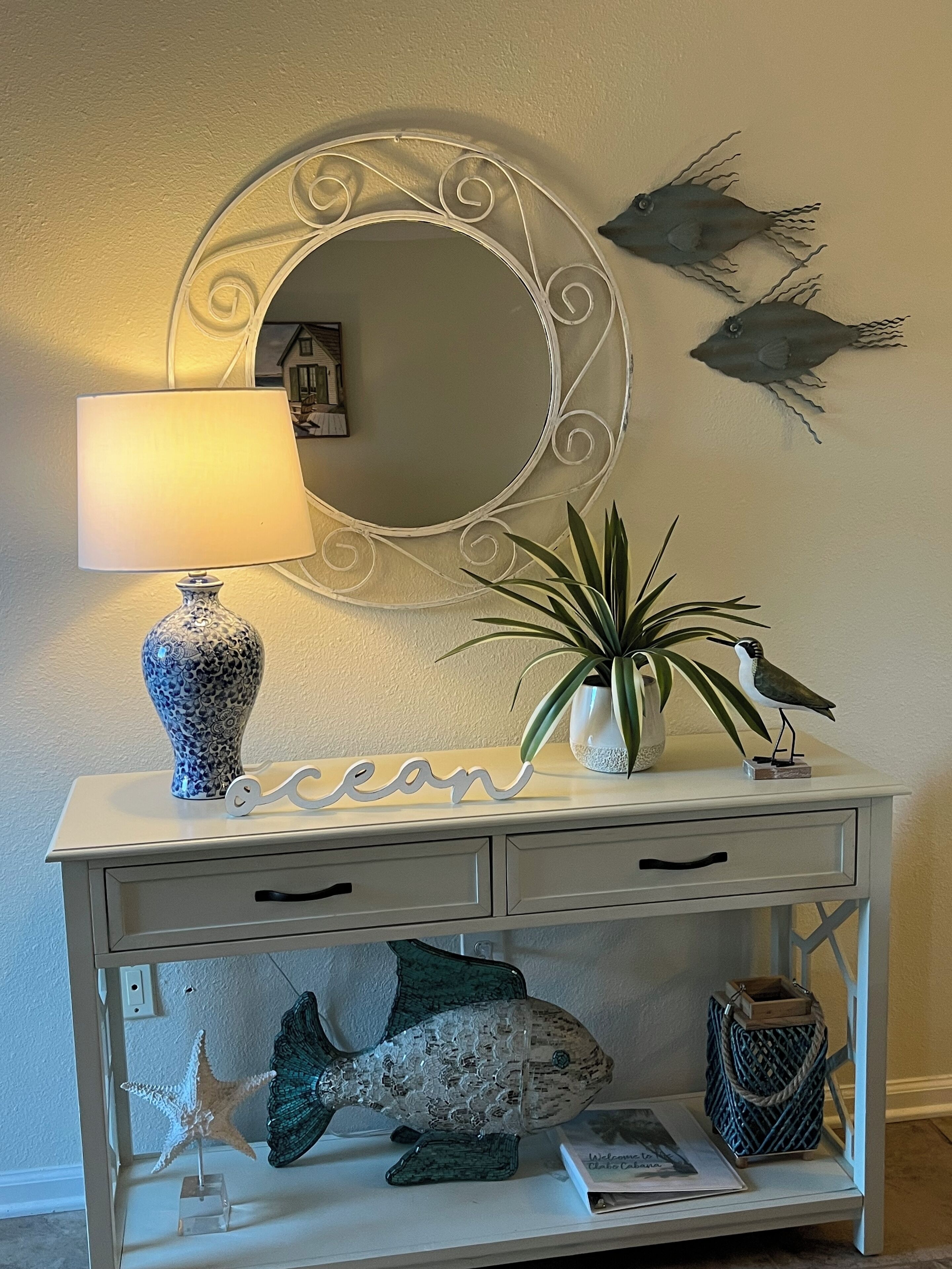 A white console table with a decorative mirror, a lamp, a potted plant, and a wall-mounted fish decoration.