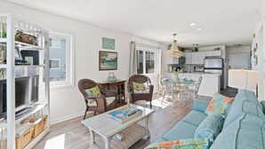 A spacious living area with a sofa, chairs, a coffee table, and a television. There is a kitchen in the background with a refrigerator and a dining table.