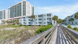 A coastal hotel with multiple-story buildings, a wooden boardwalk, and beachfront vegetation.