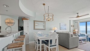 A modern kitchen with a dining area, a gray sofa, and a large window offering a view. - Beachwalk Villas 104-3BR/3BA Condo in Cherry Grove (North Myrtle Beach)