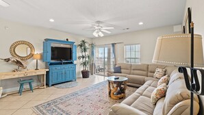 Un salón con un sofá color beige, un mueble de entretenimiento azul, un ventilador de techo y un ventanal con persianas.
