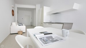 A modern, minimalist room with a white sofa, a dining table with a magazine and a mug, and a kitchen area with white cabinets and a stainless steel sink. - Rivazzurra Homes - 3 (Termoli)