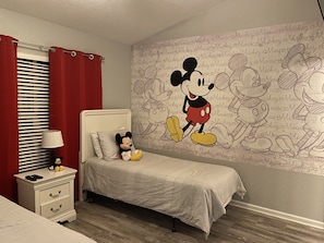 Une chambre avec une fresque représentant Mickey Mouse sur le mur, un lit avec une couvre-lit grise, une table de chevet avec une lampe et un doudou à l’effigie de Mickey Mouse sur le lit.