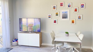 A modern living room with a flat-screen TV, a white cabinet, a dining table with chairs, and a wall decorated with travel posters.