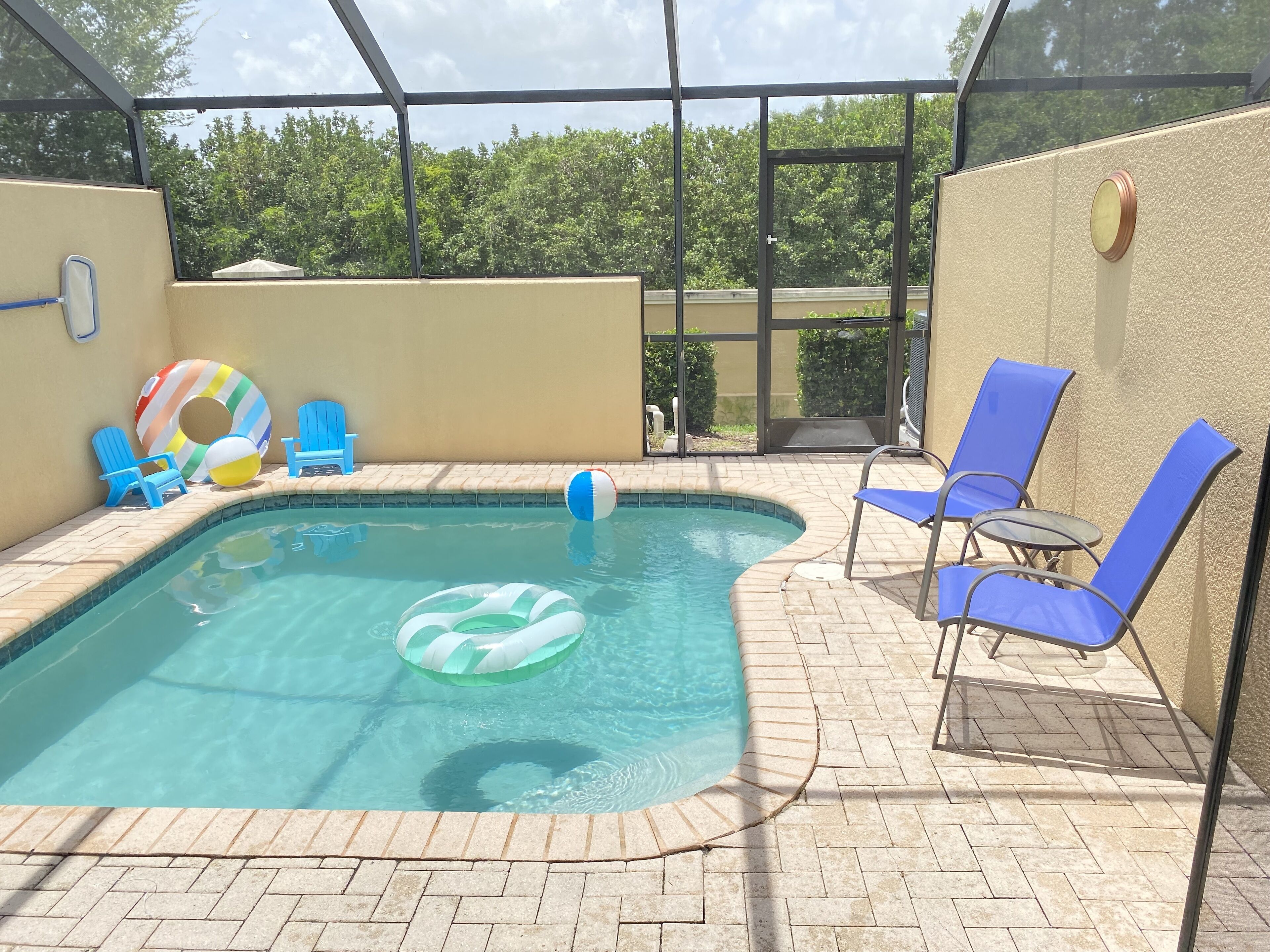 A swimming pool with blue chairs and inflatable rings, surrounded by a brick patio and glass walls.