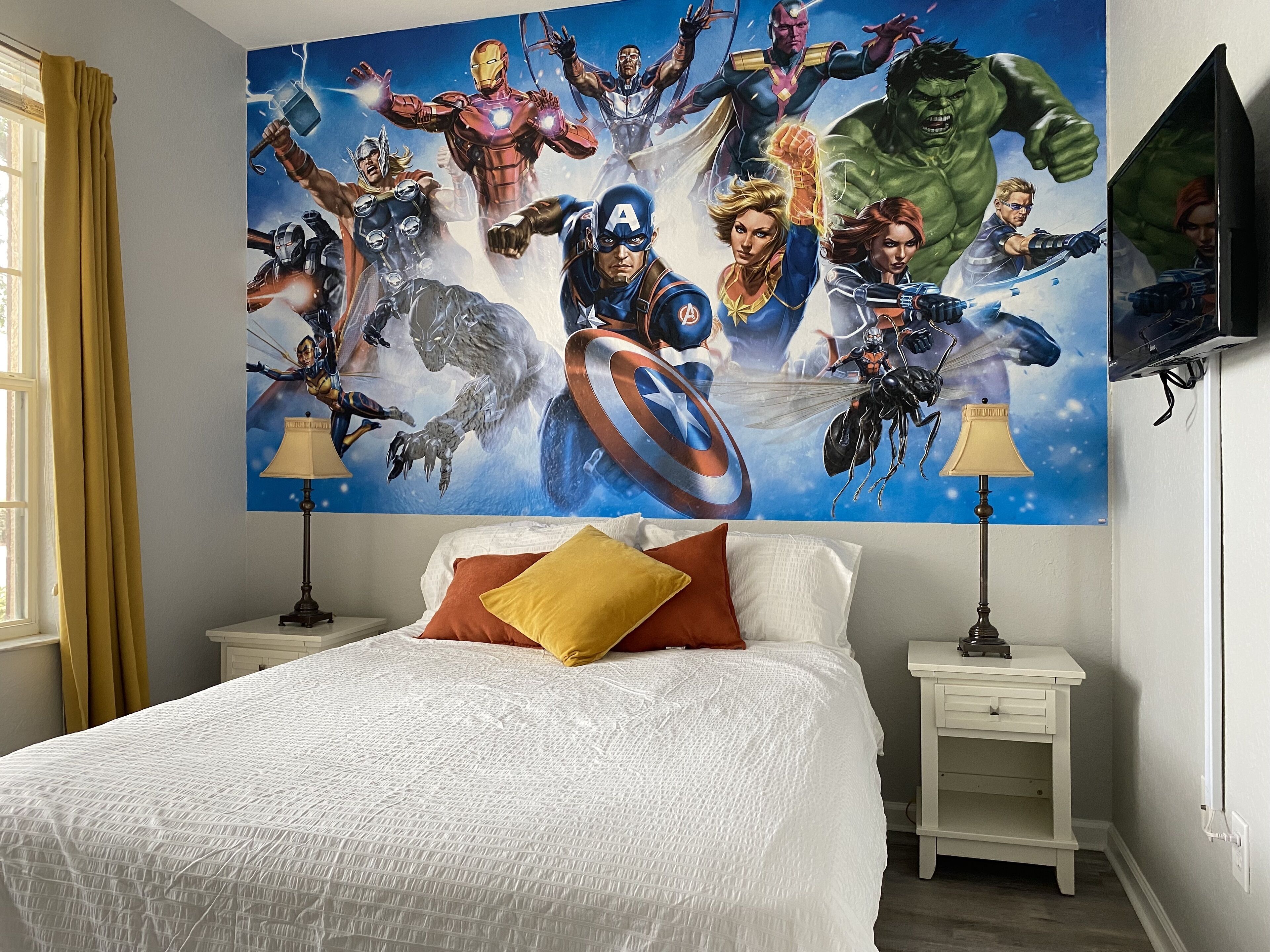 Une chambre avec une grande fresque murale sur le thème des super-héros, un lit avec une literie blanche, deux tables de chevet avec des lampes et une fenêtre avec des rideaux.