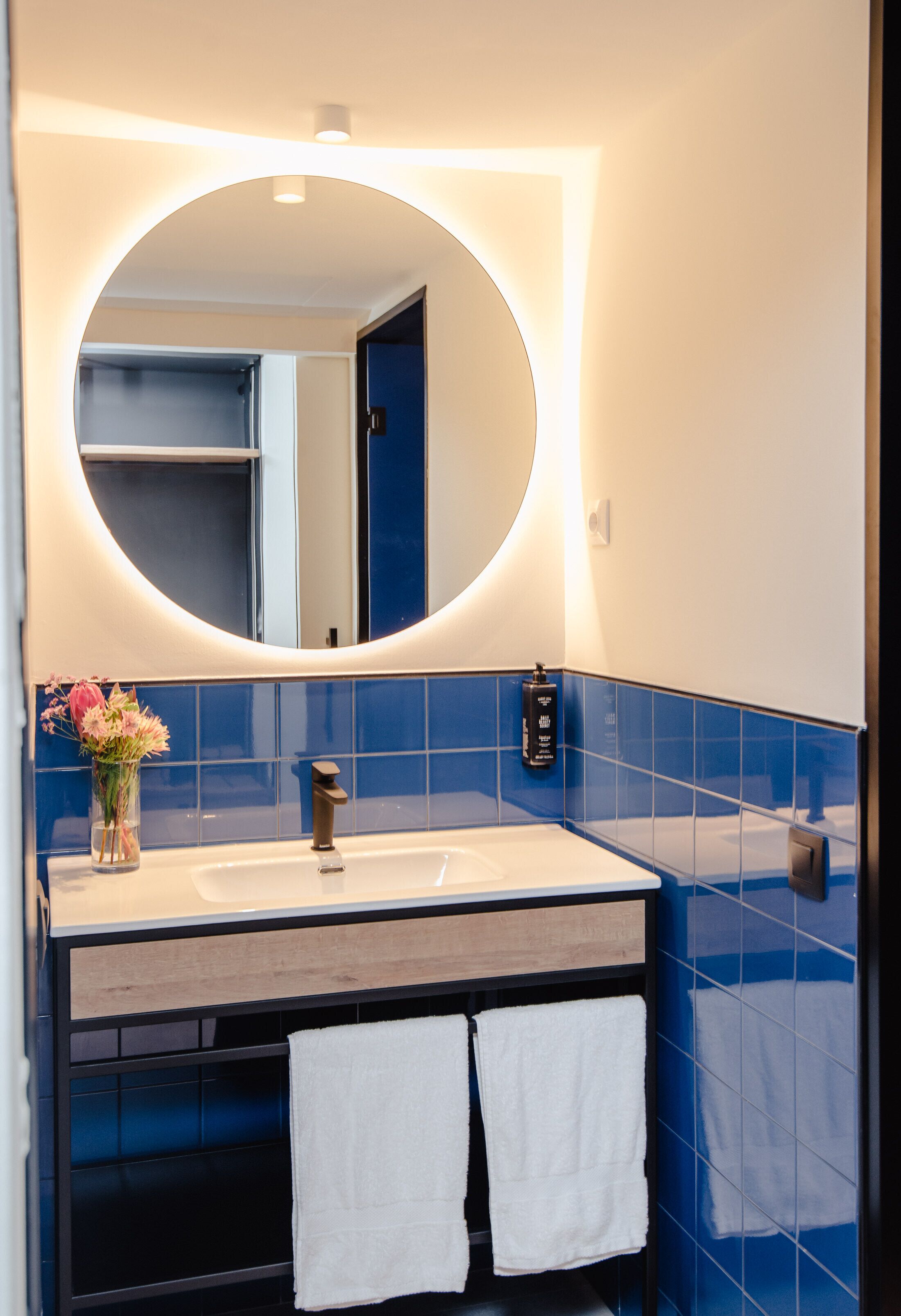 Un baño con un espejo redondo, un lavamanos blanco, azulejos azules y dos toallas blancas.