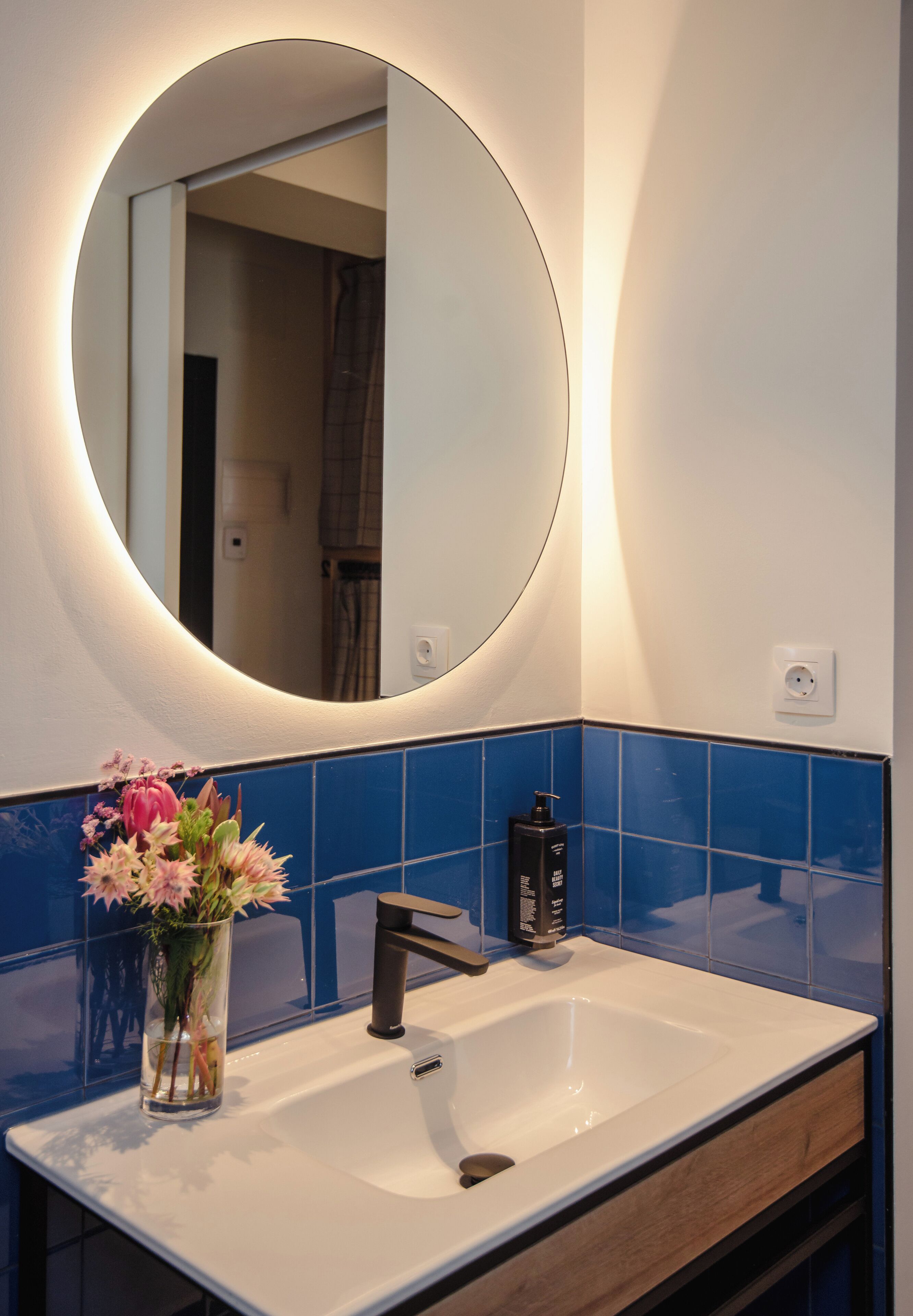 A bathroom with a round mirror, a white sink, blue tiles, a wooden cabinet, and a vase with flowers.