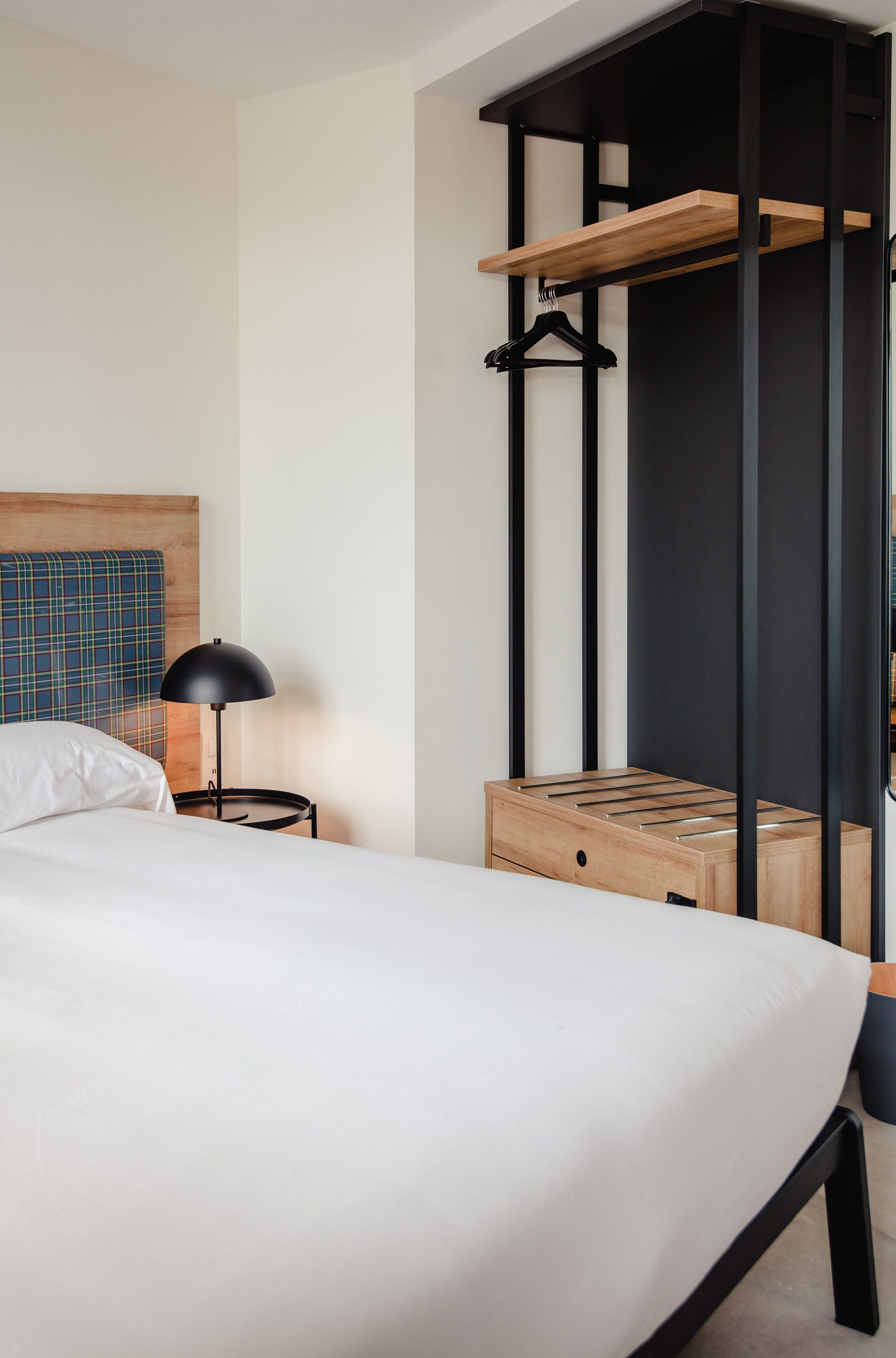 A modern hotel room with a bed, a bedside table with a lamp, a wooden headboard, a black metal shelf, and a small wooden chest.