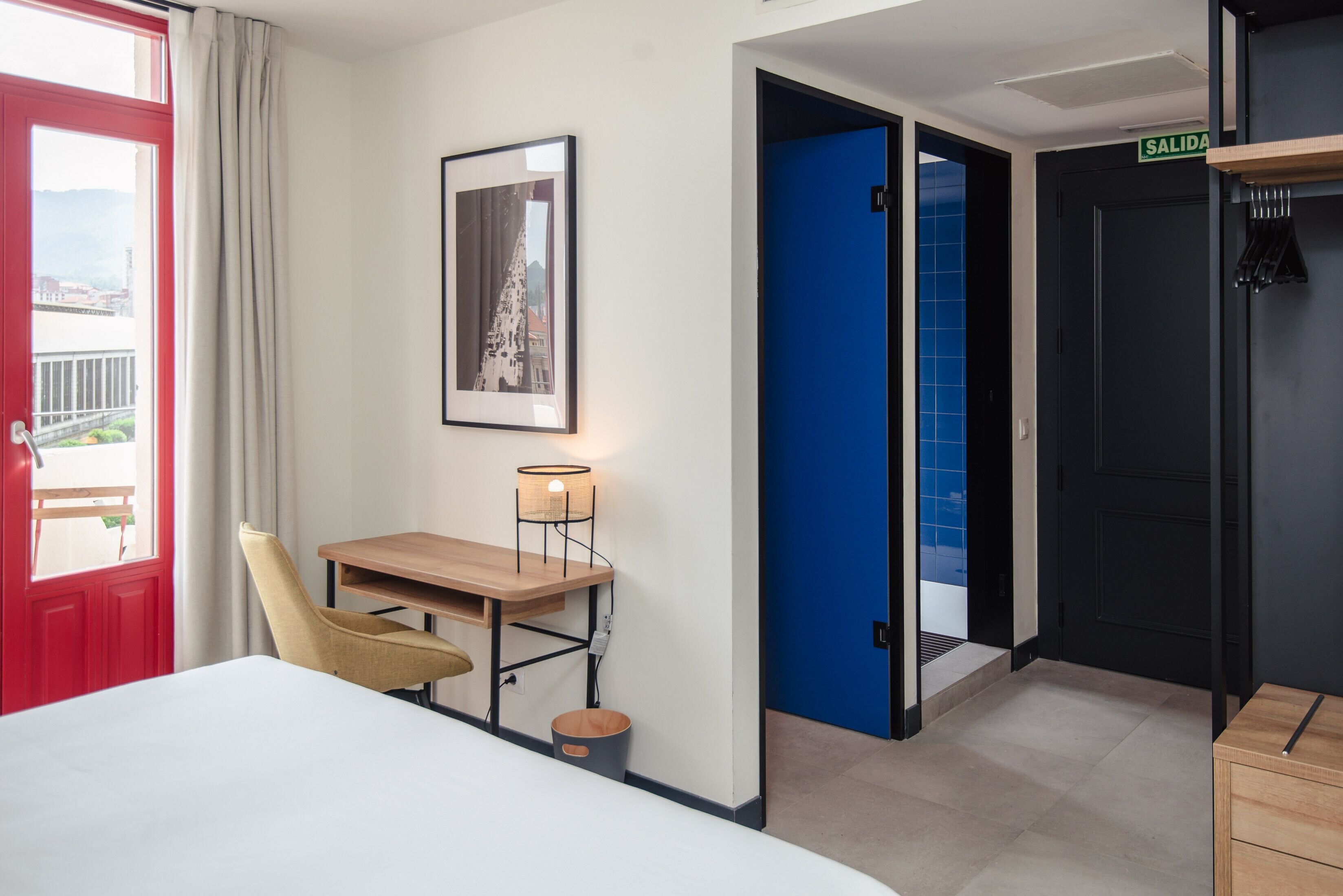 Habitació d'hotel amb un llit, una taula de treball amb una cadira, un quadre al mur, una làmpada i una porta que dóna a un bany.