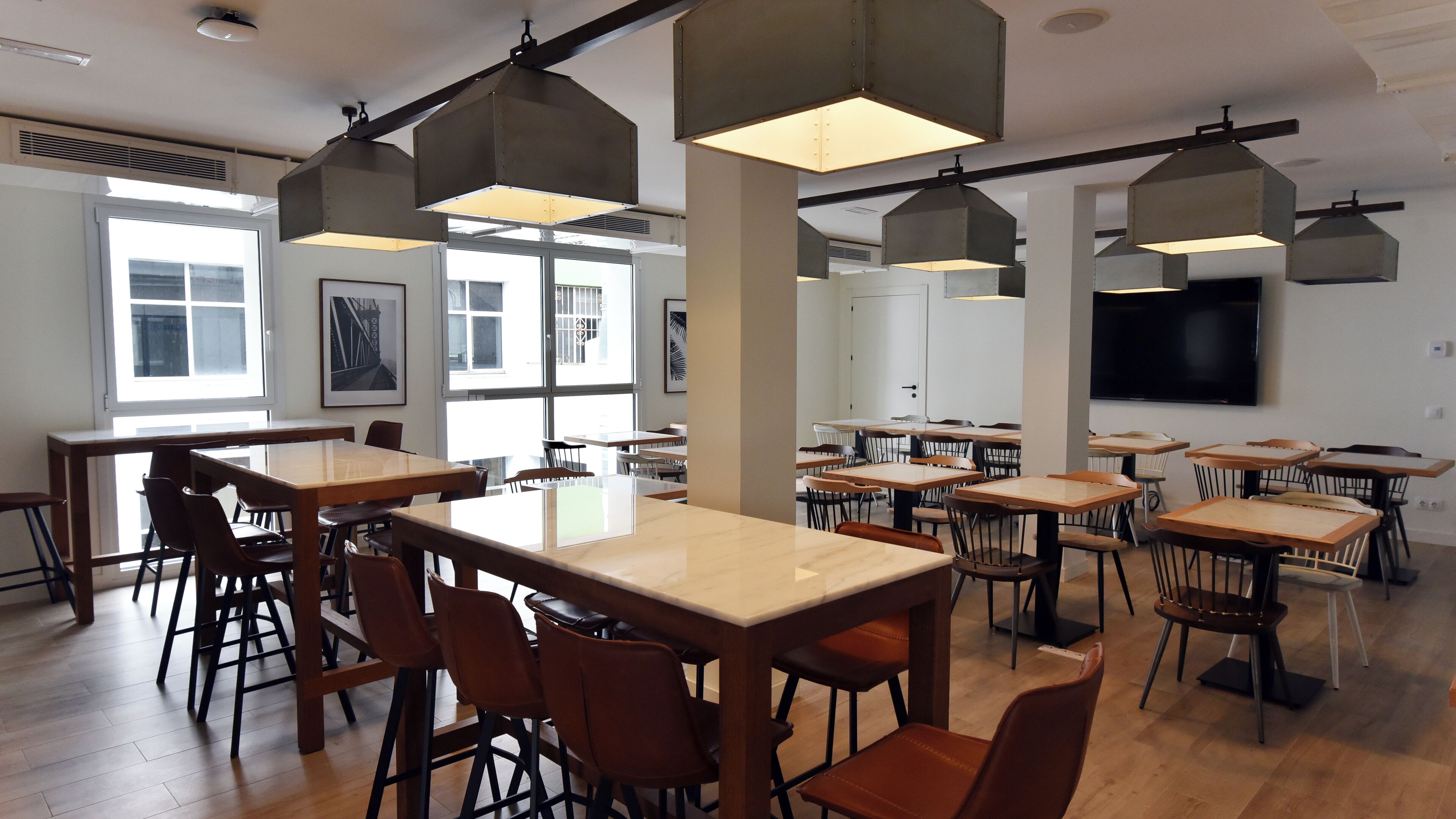 Een ruime eetruimte met hoge tafels en stoelen, grote ramen en hangende lampen in industriële stijl.