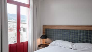 Habitación de hotel con cama, mesita de noche, lámpara, ventana con cortinas y vistas a edificios y colinas.
