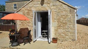Ein Steinhaus mit geöffneter Tür, einem Tisch mit Stühlen und einem orangefarbenen Sonnenschirm.