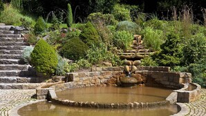 Ein Steinkunstbrunnen, überzogen mit Wasserfällen, umgeben von einem kreisförmigen Steinweg und üppiger Vegetation.