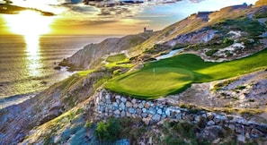 Un parcours de golf situé sur la côte, avec un mur de pierre, un drapeau et un bâtiment visible au loin.