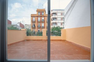 Vista a través de una ventana que muestra un balcón con piso de baldosas, baranda de metal y un edificio con balcones al fondo.