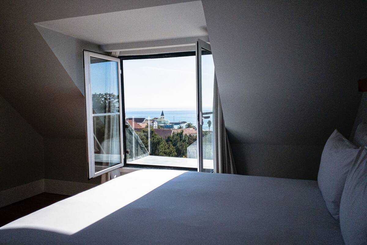 A bedroom with a large window, a bed, and a view of the sea and buildings.