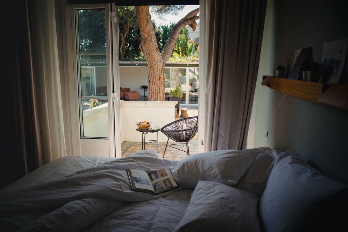 A bedroom with a bed, an open door leading to a balcony with a chair and table, and a tree outside.