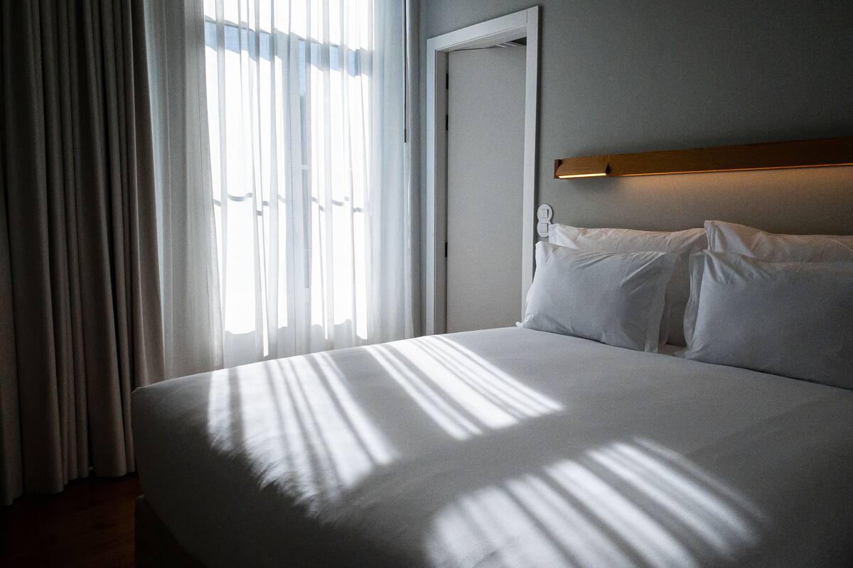 A neatly made bed with white linens, a wooden headboard, and a wall-mounted light fixture.