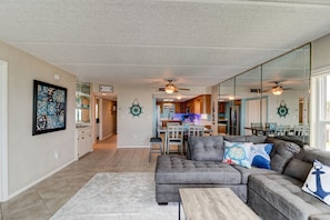 A spacious living room with a gray sectional sofa, a wooden coffee table, and a large mirror reflecting the kitchen area. - Oceanfront Sunrise 2 Bed 2 Bath Condo with Pool (Ormond Beach)